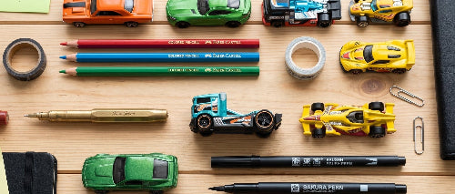 A styled flat-lay of miniature toy race cars arranged on a light wood desk alongside stationery items like pens, sticky notes, paper clips, and a black notebook.