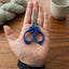 Small scissors with bright blue handles and silver metal blades resting on an open hand, photographed over a wooden tabletop with yarn and a ceramic cup in the background.
